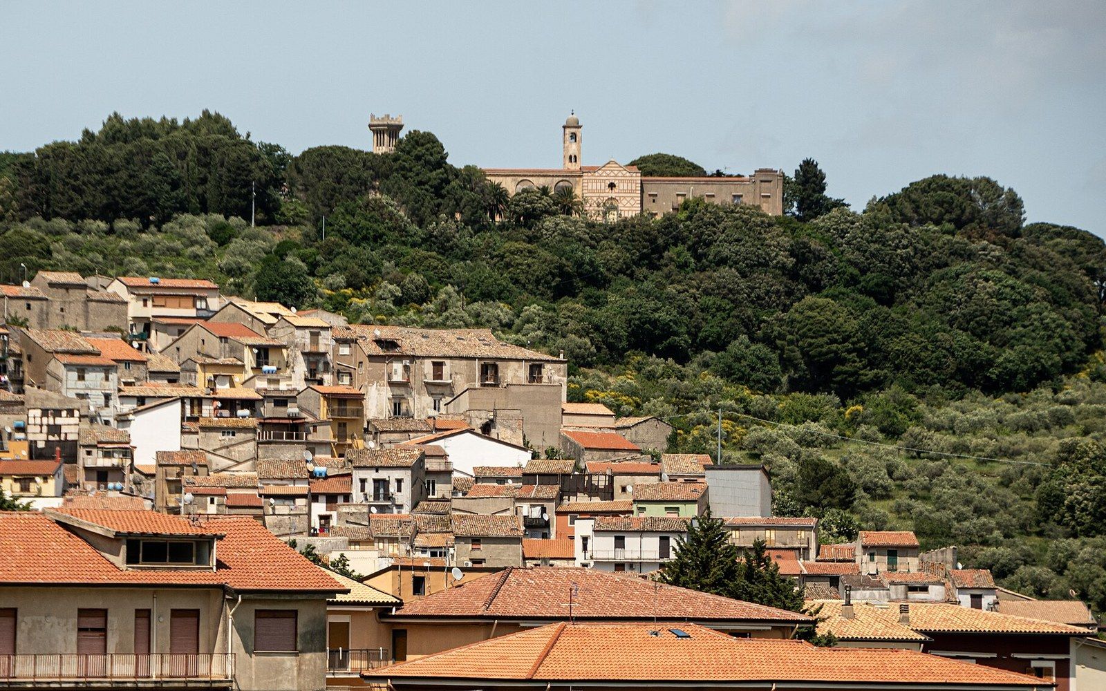 A town of tiled roofs below a wooded hill topped by a church and tower
