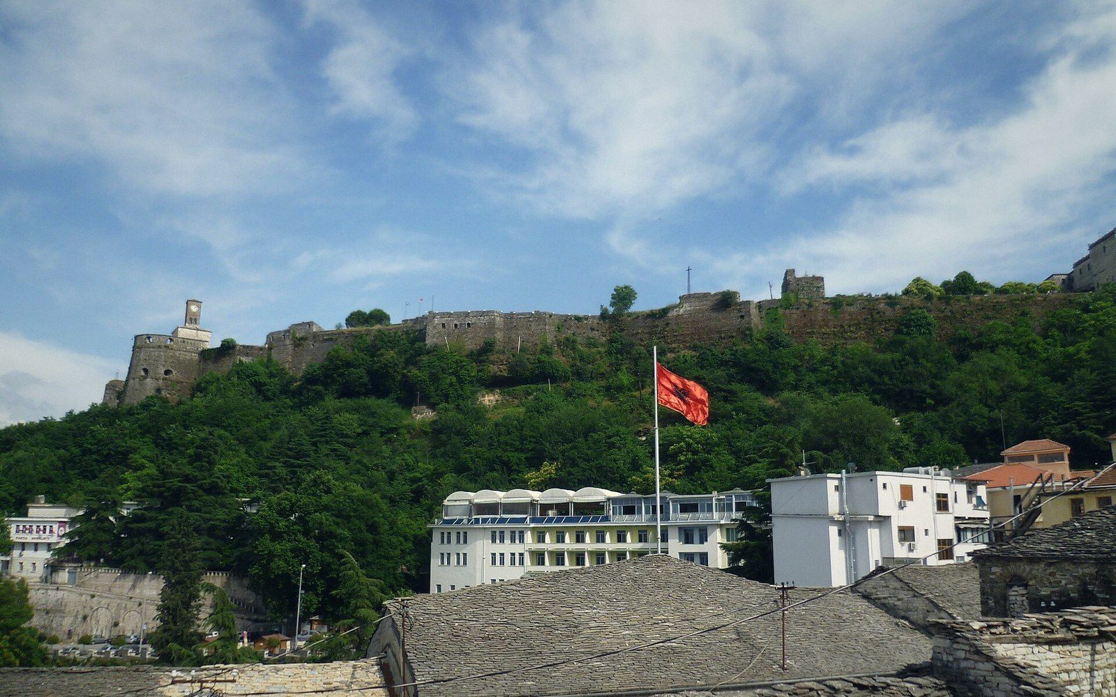 A hilltop castle and clock tower above a town of grey slate roofs, an Albanian flag flying