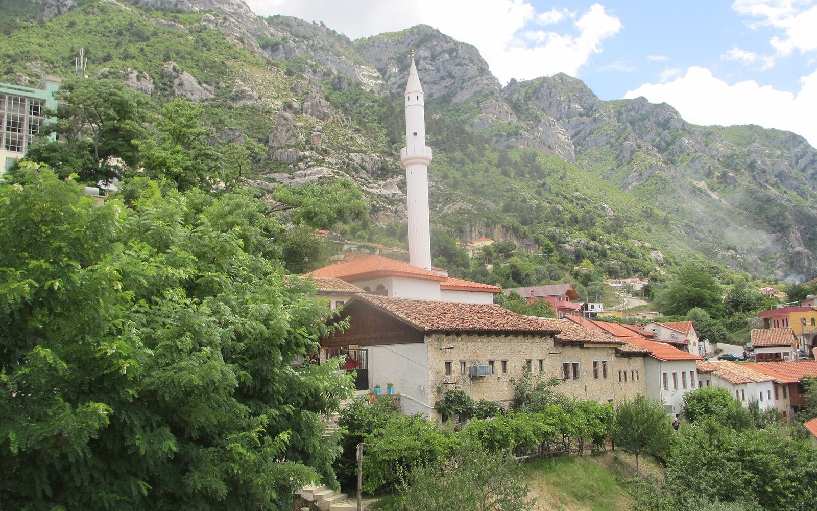 Timber bazaar shops and a white minaret below a mountain wall under a storm sky